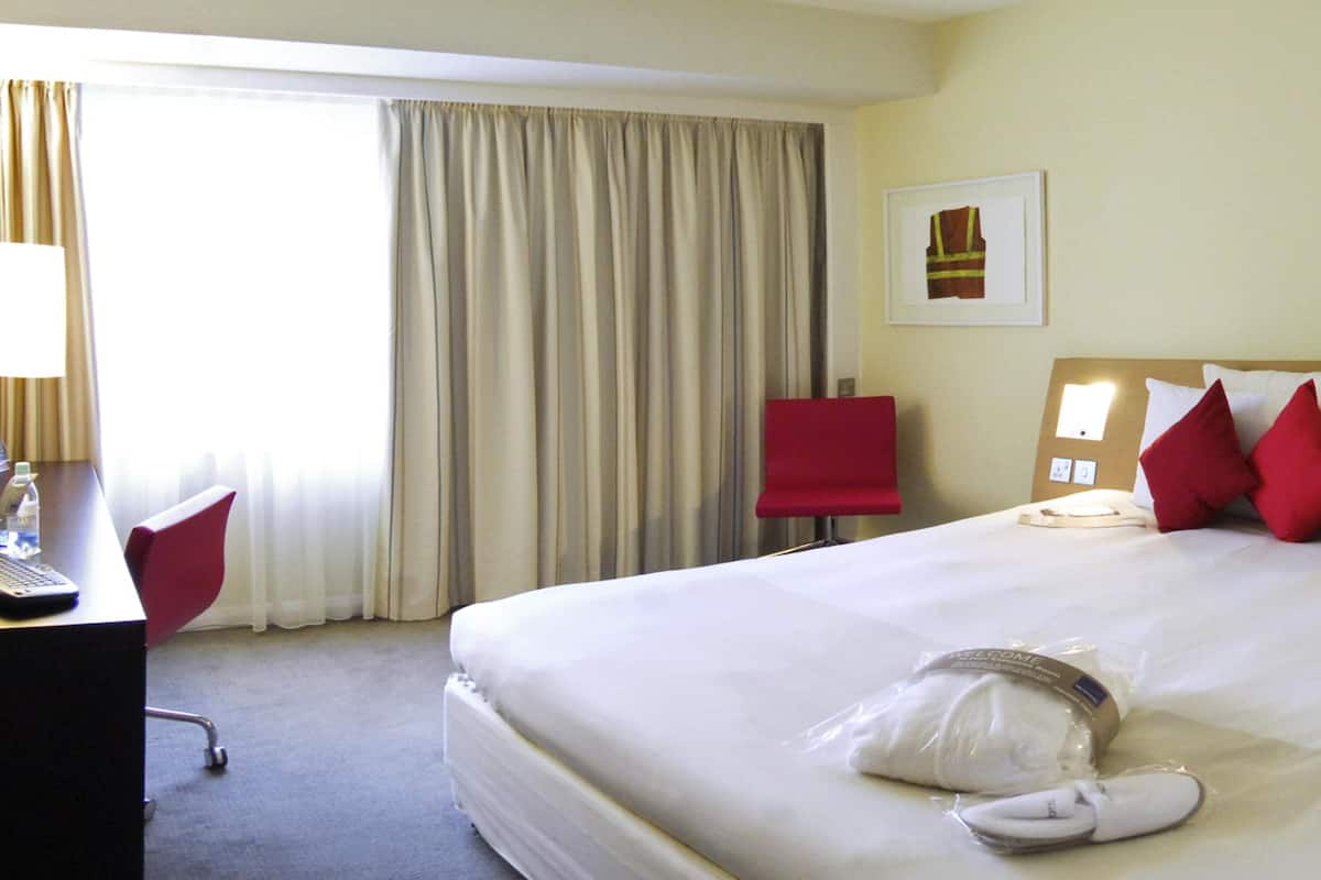 A hotel room with a large bed, a desk with a computer, a chair, and a window with curtains.