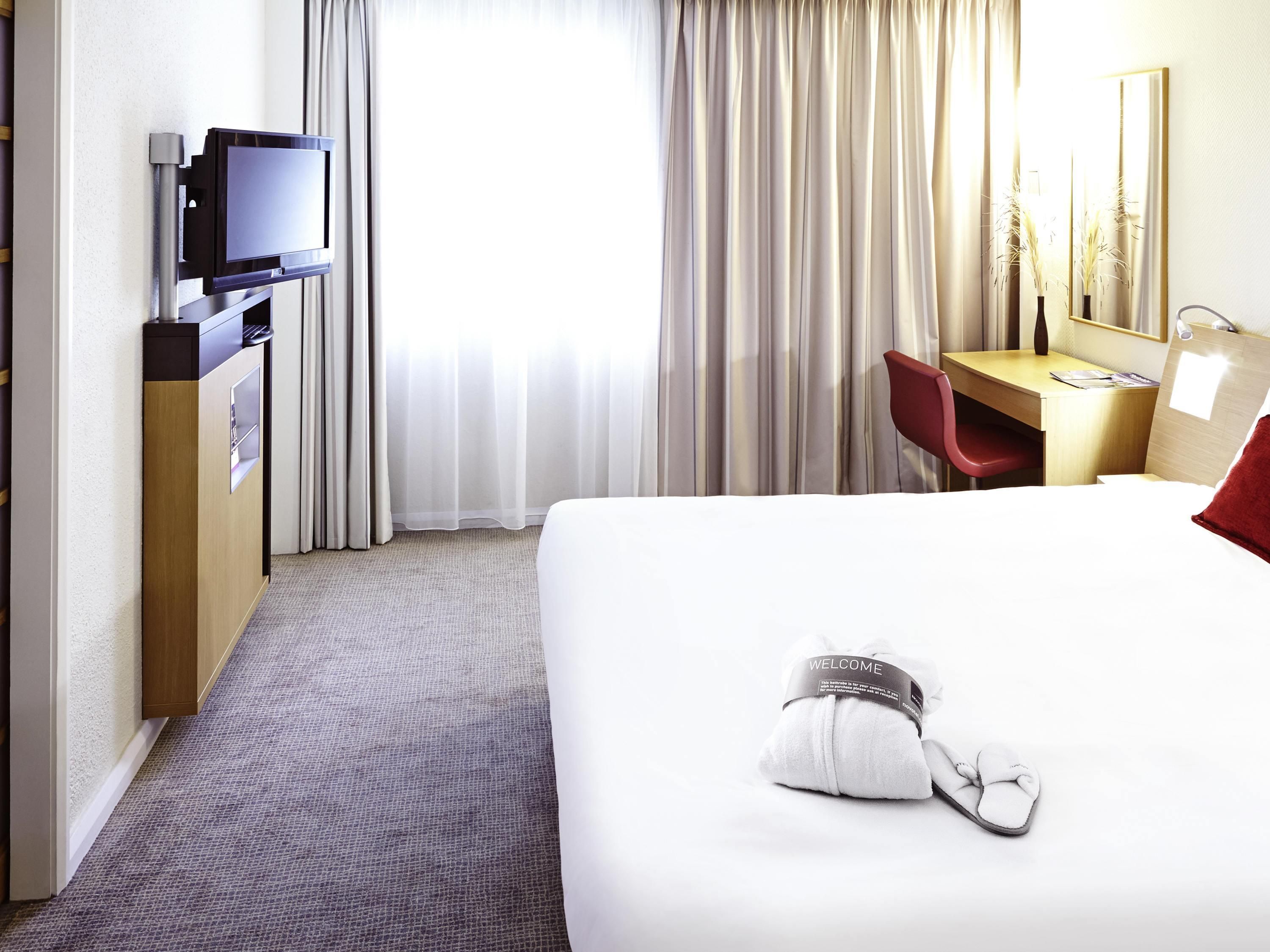 Quarto de hotel com cama, escrivaninha com cadeira, televisão e janela com cortinas.