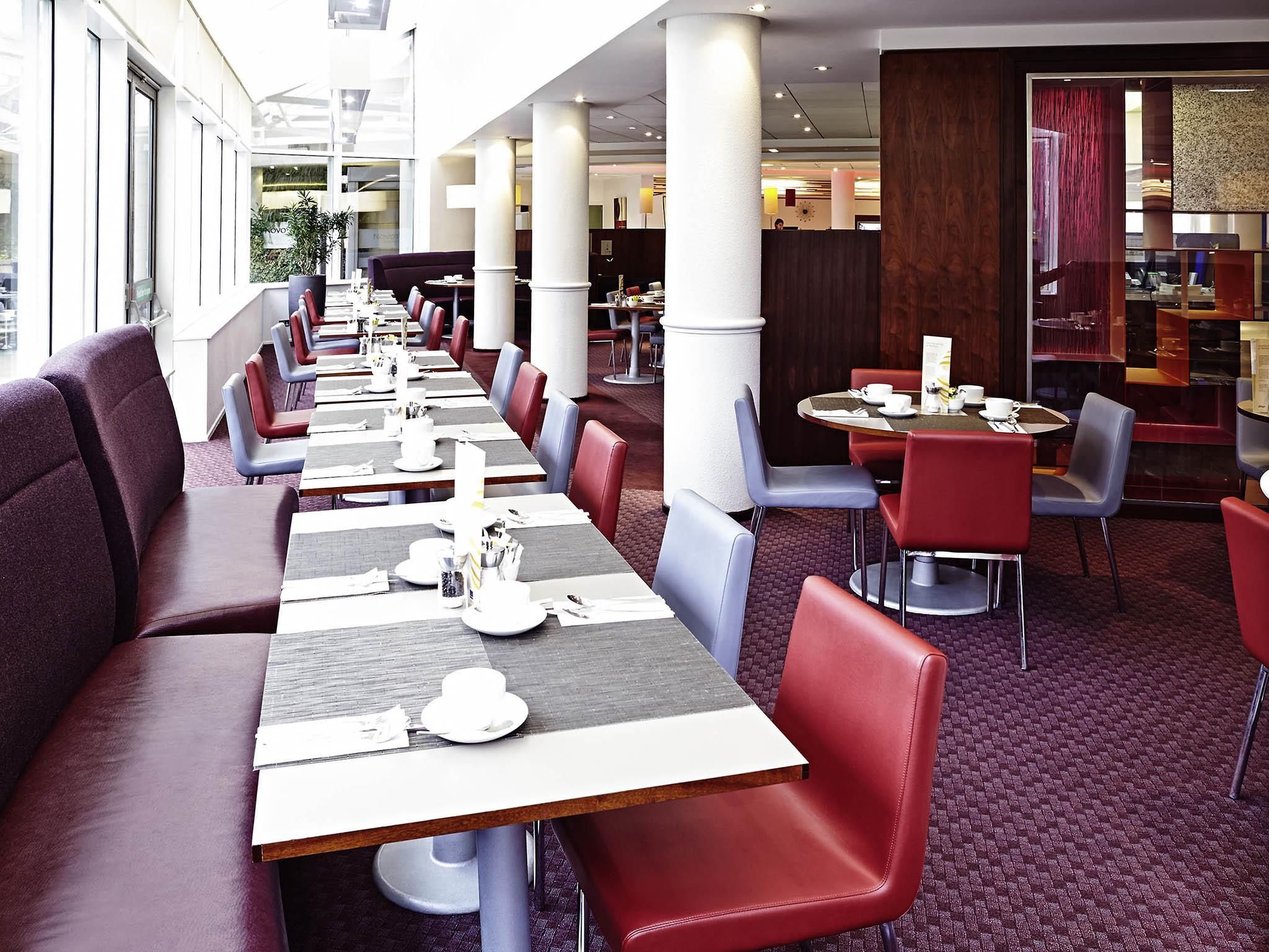 A spacious restaurant with long tables and red and grey chairs, featuring a modern interior and large windows.