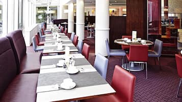 A spacious restaurant with long tables and red and grey chairs, featuring a modern interior and large windows.