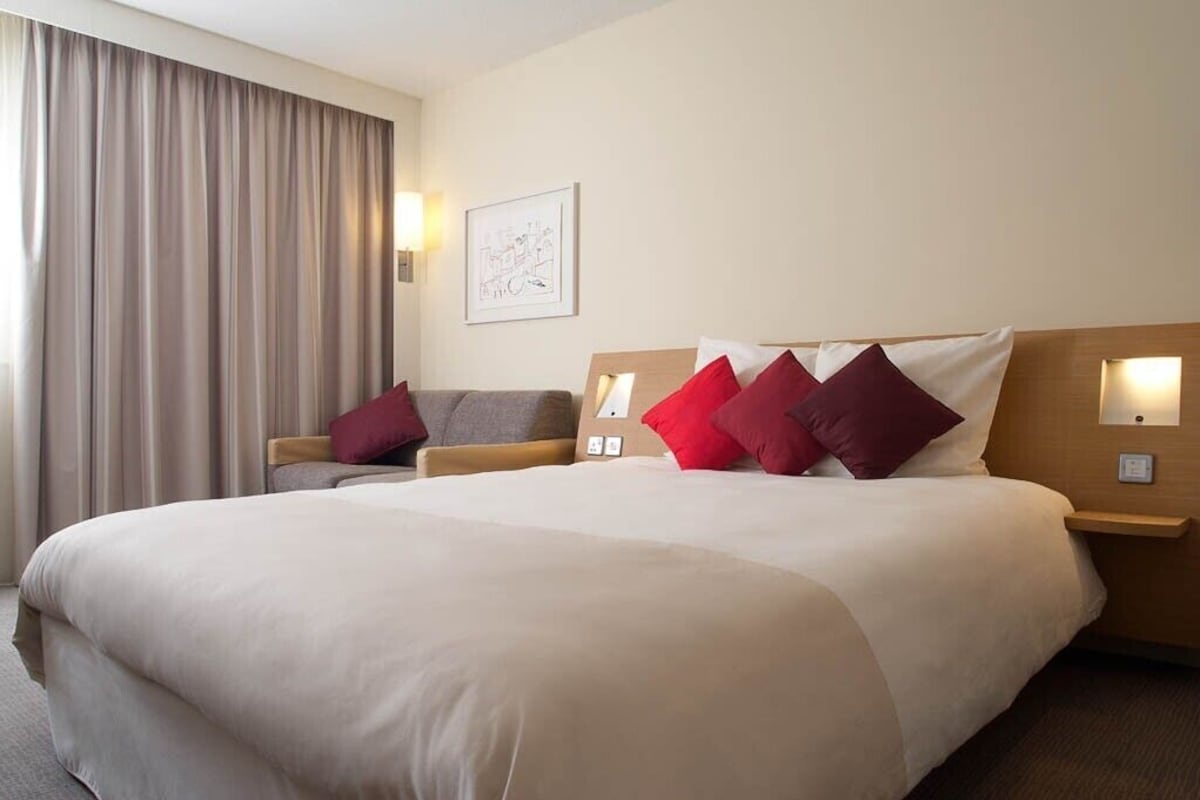 A hotel room with a large bed, two bedside tables with lamps, a sofa, and a large window with curtains.