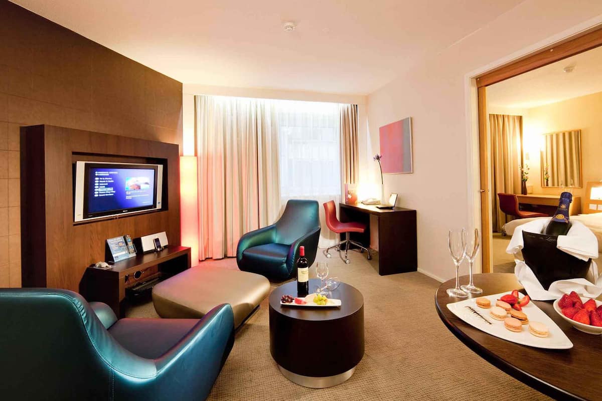 A hotel room with a TV, comfortable seating, a desk, and a dining area with snacks.