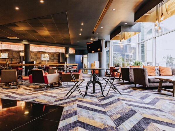 Un hall d’hôtel moderne doté d’un bar, de différents espaces aménagés pour s’asseoir et de grandes fenêtres.
