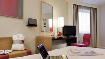 A hotel room with a bed, a desk with a laptop, a TV, a red chair, a mirror, and a window with curtains.