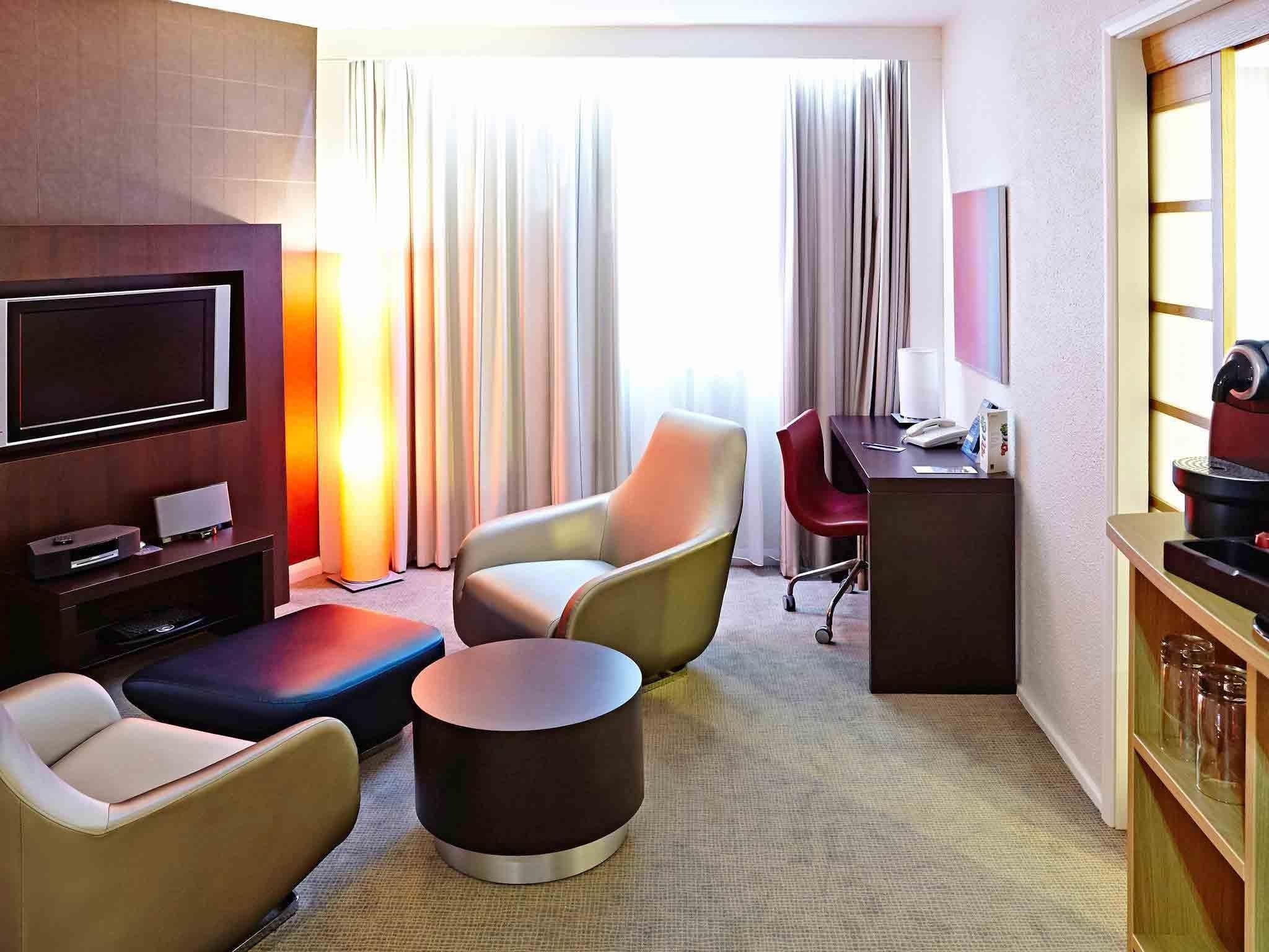 Une chambre d’hôtel équipée d’un téléviseur à écran plat, d’un bureau avec un ordinateur, d’une machine à café et d’un coin salon comprenant un canapé et des fauteuils.