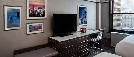 A hotel room with a bed, a desk with a flat-screen TV, a bench, and wall-mounted artwork.