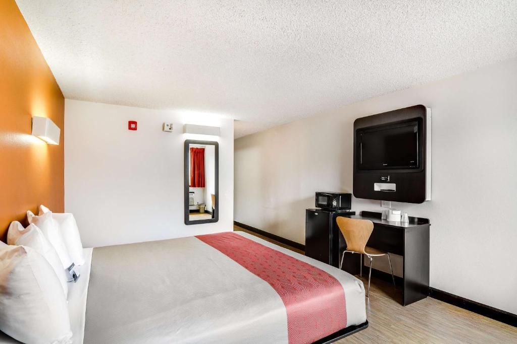 Habitación de hotel con cama, escritorio, silla, televisor y espejo.