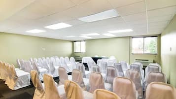 A conference room with rows of chairs covered in white and beige chair covers, a table with a white tablecloth, and a window with blinds.
