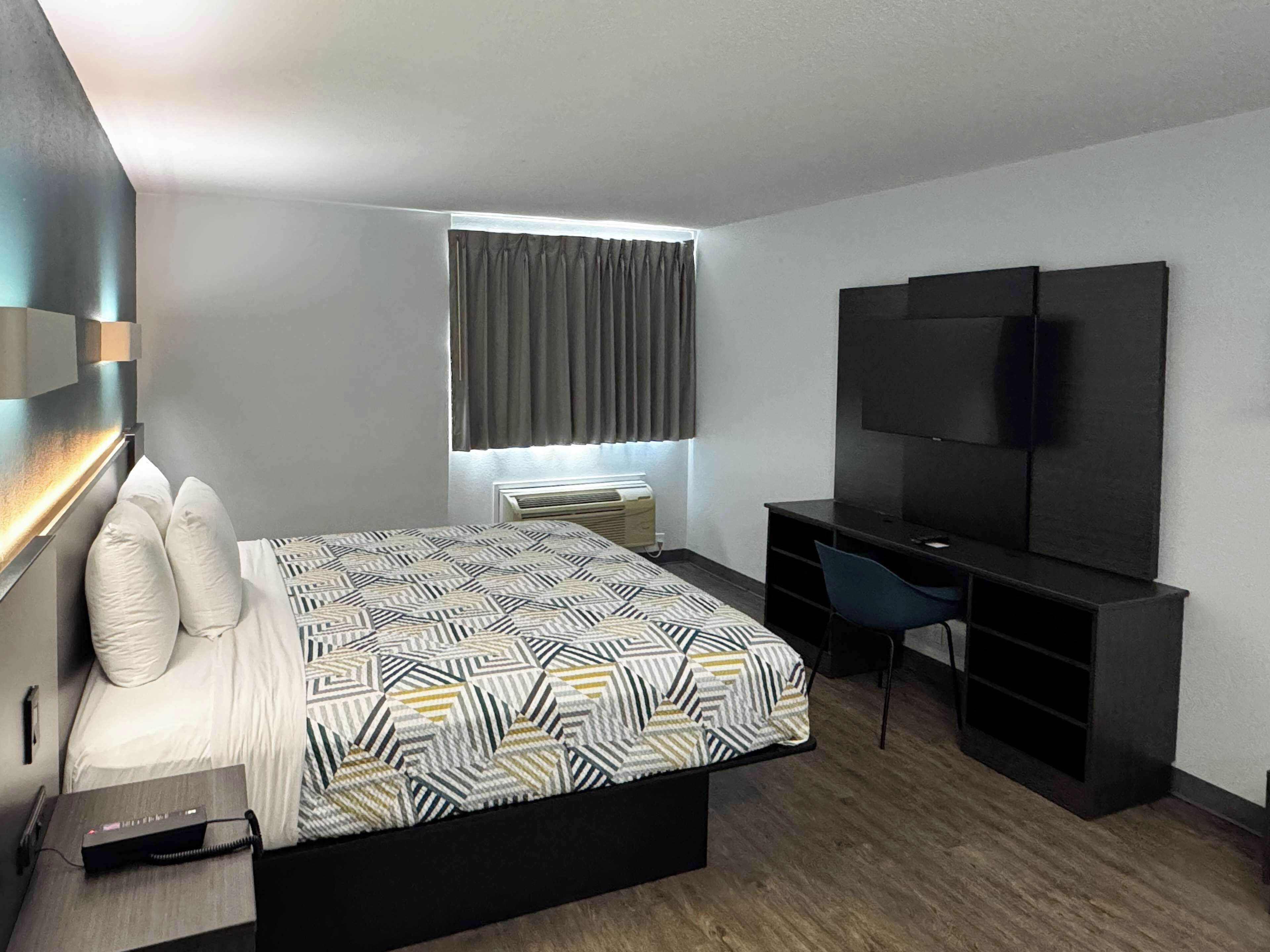 A modern hotel room with a bed, a desk, a chair, a TV, and a window with curtains.