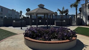 A gazebo with a tiled roof, surrounded by a brick planter with purple flowers, and a gated entrance with palm trees.