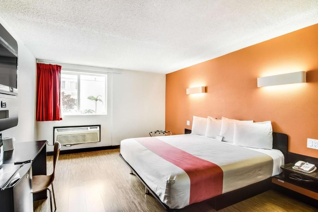 Une chambre d’hôtel comprenant un lit, un bureau, une chaise, une fenêtre avec des rideaux, un radiateur fixé au mur et une table de chevet avec un téléphone.