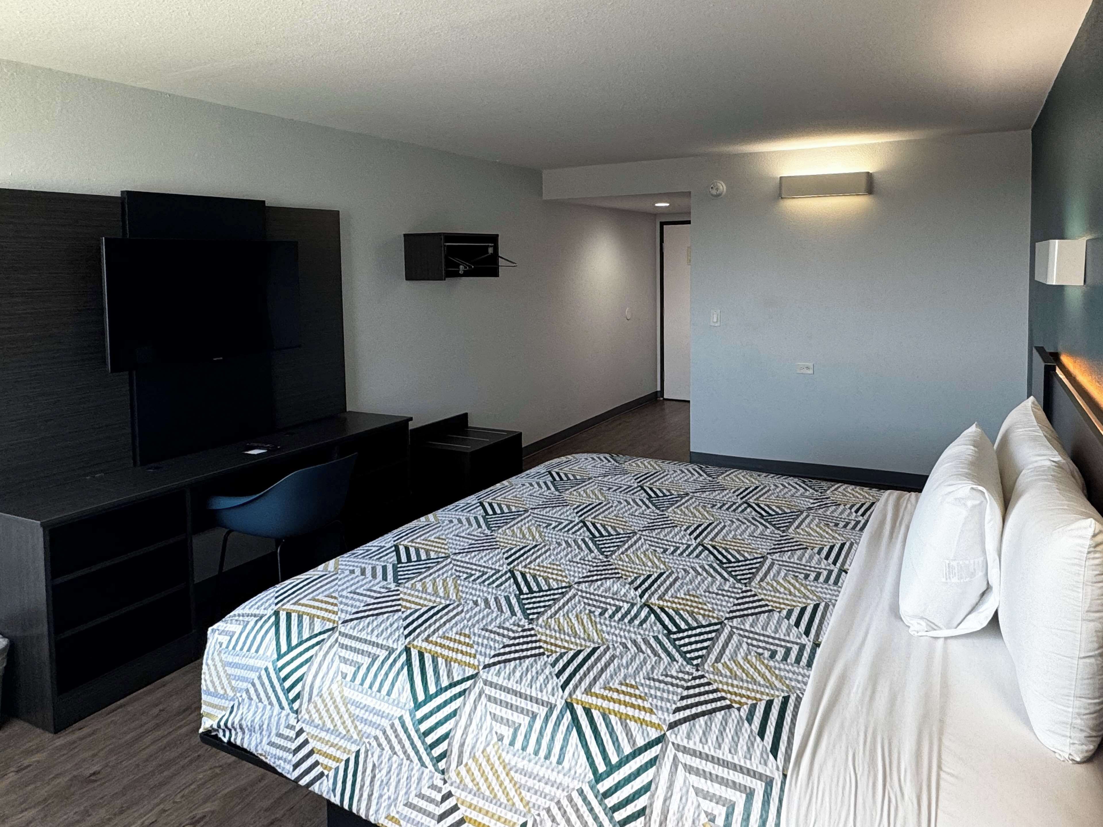 Una habitación de hotel moderna con una cama grande, un escritorio con silla, un televisor de pantalla plana y un aplique de pared.