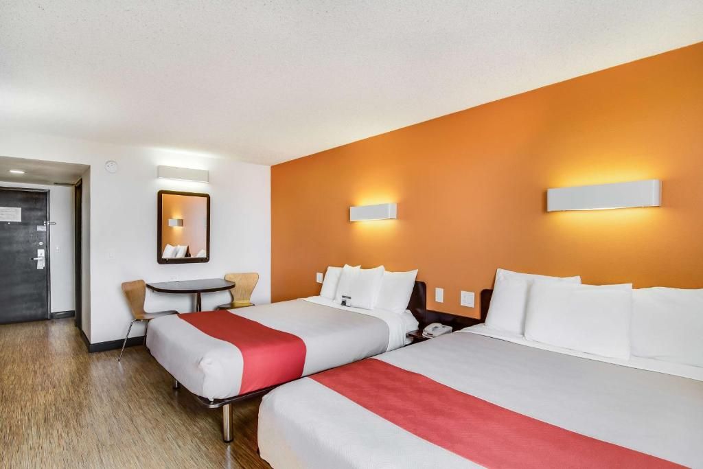 A hotel room with two beds, a small table, a chair, and a door.