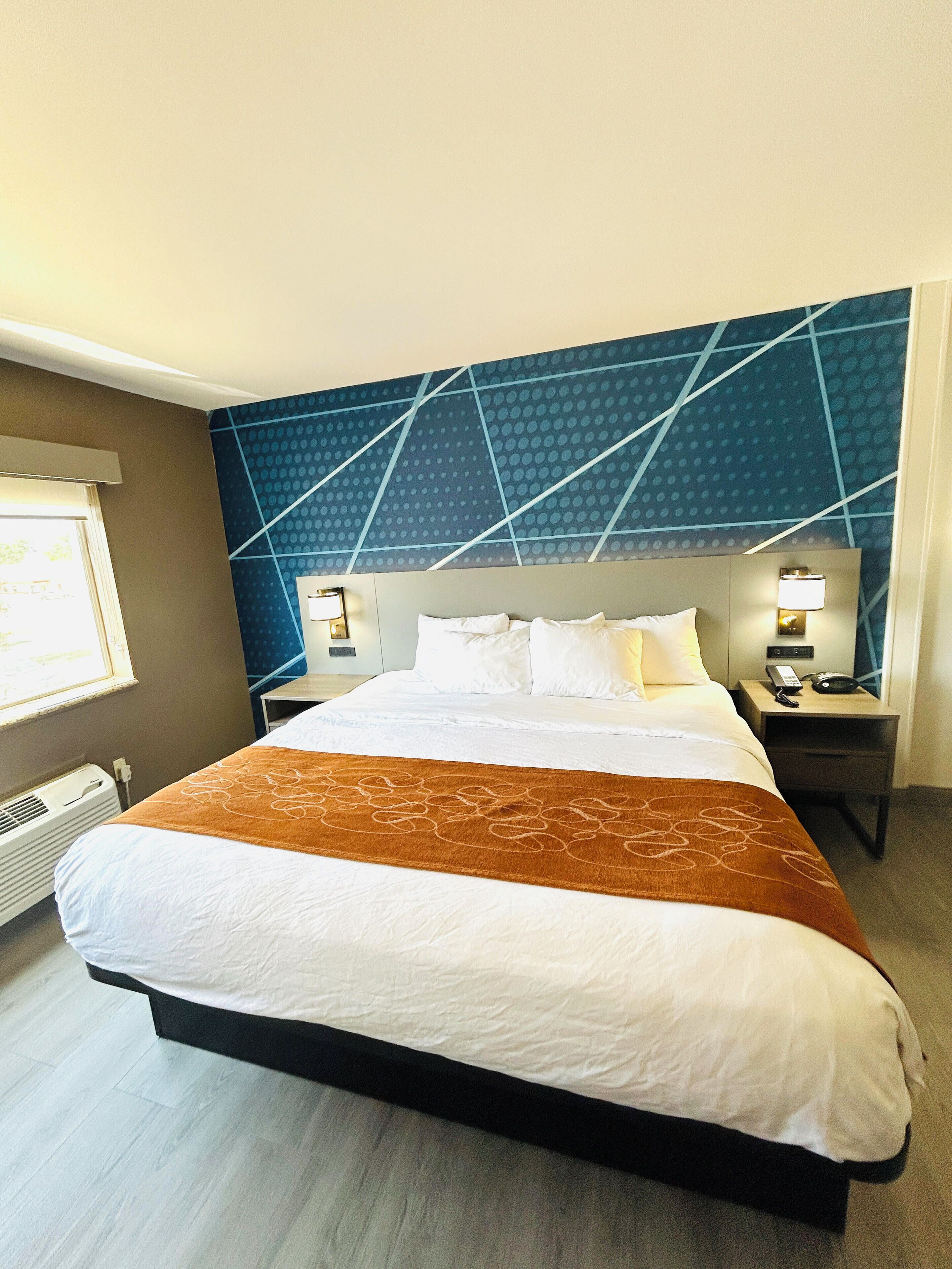 Habitación de hotel con una cama grande, cabecera con diseño geométrico, mesita de noche con lámpara y ventana con persianas.
