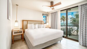 A hotel room with a large bed, a ceiling fan, a nightstand with a lamp, and a view of the beach through the window. - Chesapeake Beach Resort (Islamorada)