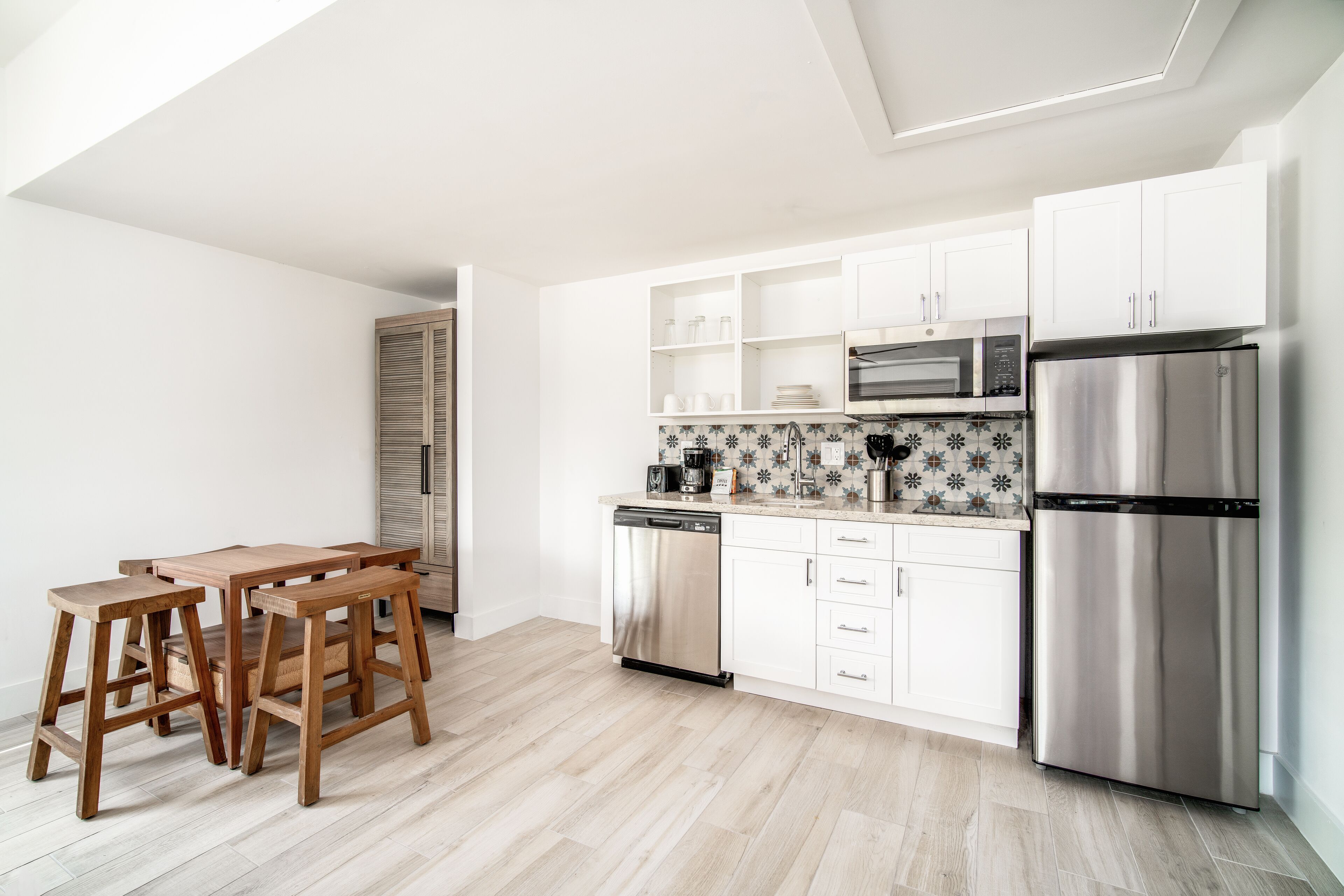 A modern kitchen with white cabinets, a stainless steel refrigerator, a microwave, and a dishwasher. There is a wooden dining table with four chairs.