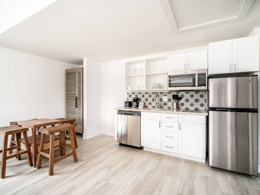 A modern kitchen with white cabinets, a stainless steel refrigerator, a microwave, and a dishwasher. There is a wooden dining table with four chairs.