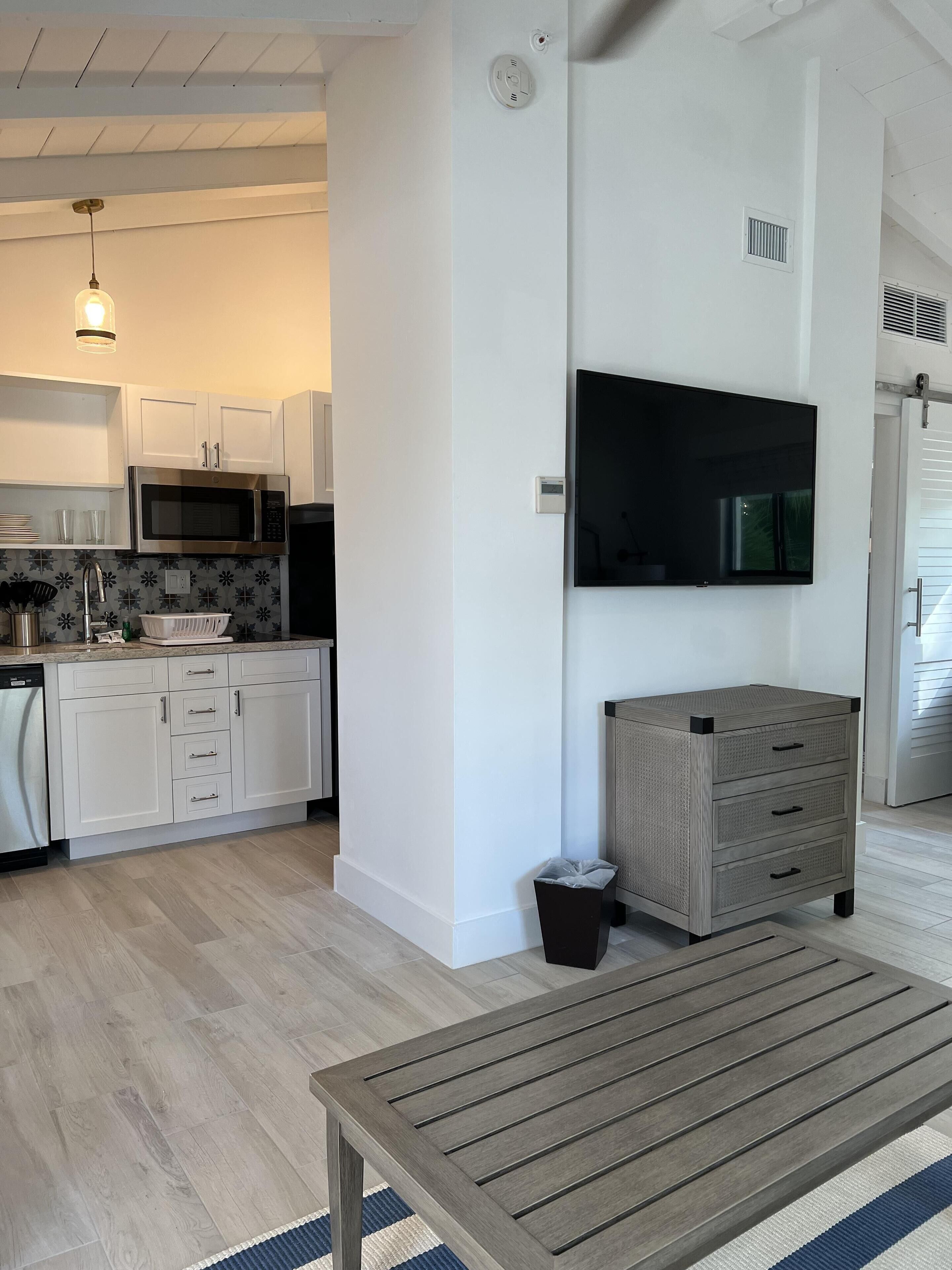 A modern kitchen with white cabinets, a microwave, and a sink. There's a flat-screen TV mounted on the wall, a small trash bin, and a wooden table with a striped rug.