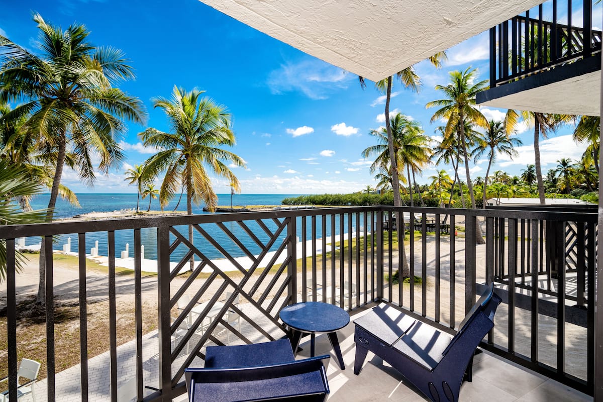 A balcony with a view of the ocean and palm trees, featuring a chair, a small table, and a bench.