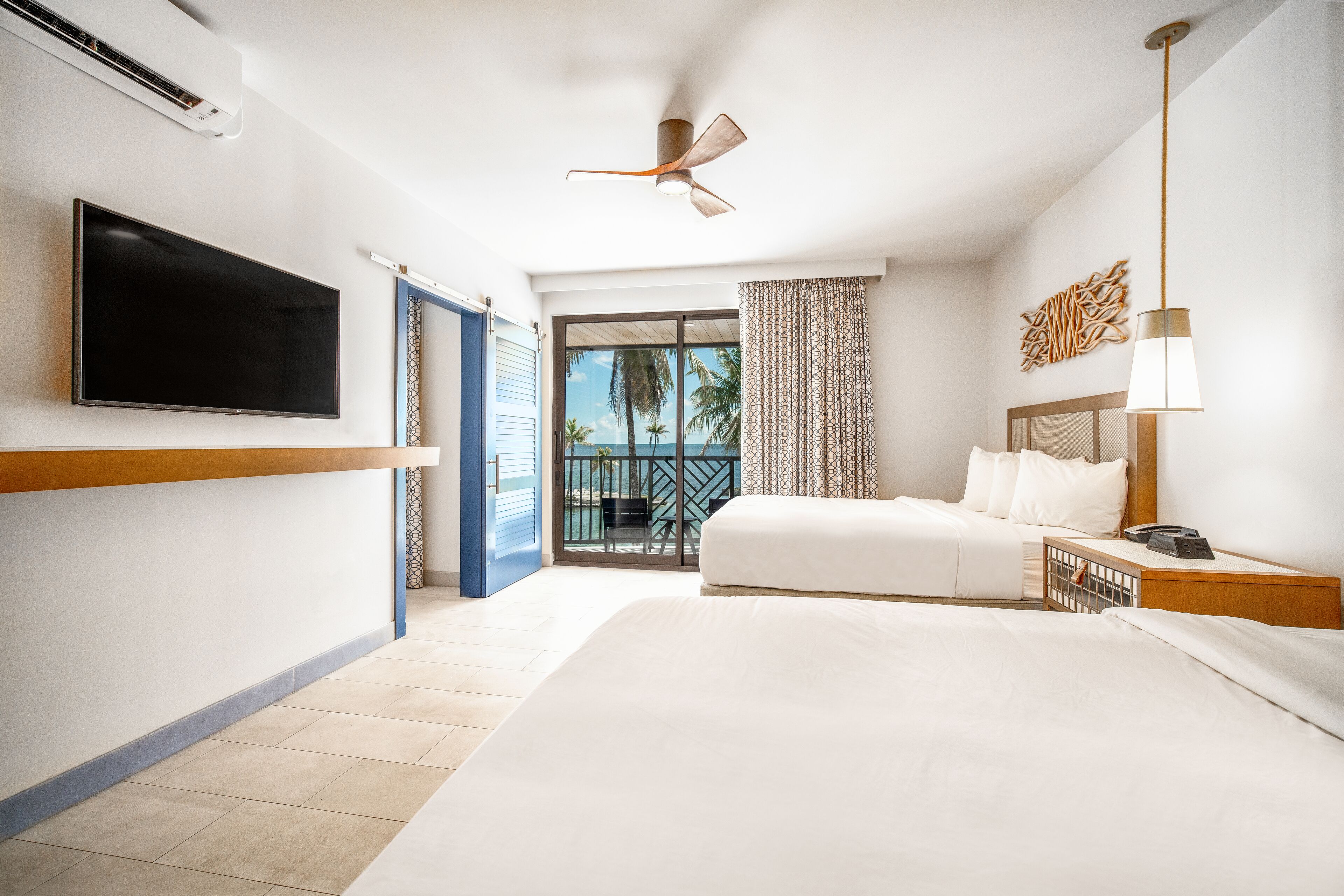 A hotel room with a large bed, a flat-screen TV, a ceiling fan, and a balcony with a view of palm trees.