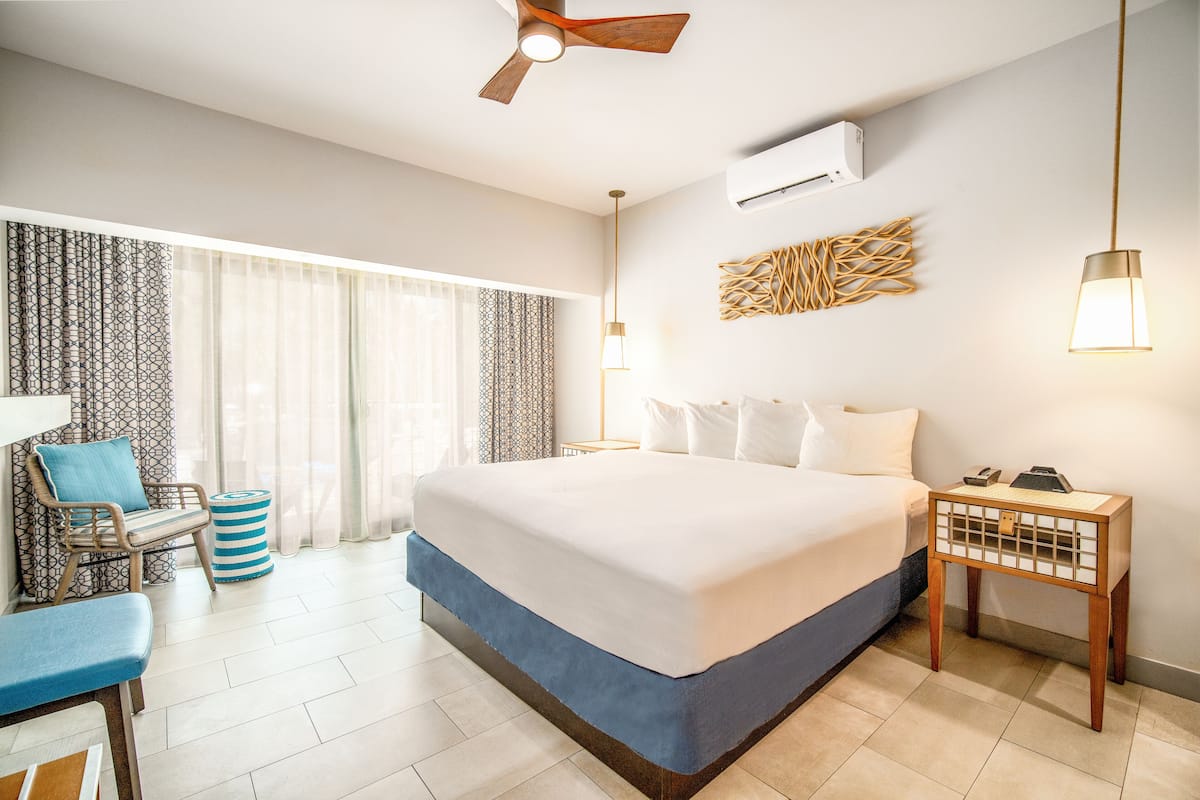A hotel room with a large bed, a desk with a chair, a ceiling fan, and an air conditioning unit.