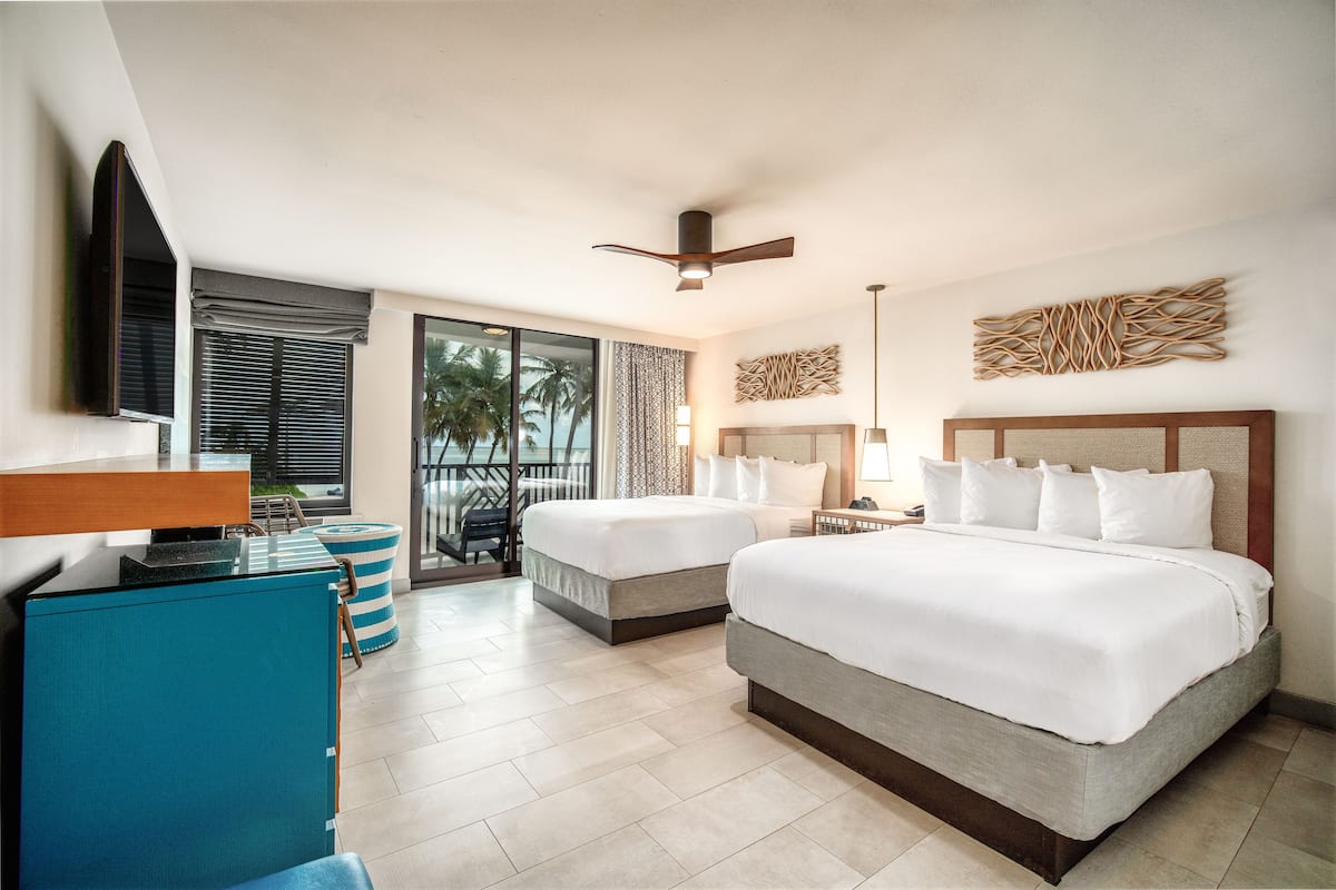 A hotel room with two beds, a desk, a TV, and a balcony with a view of palm trees.