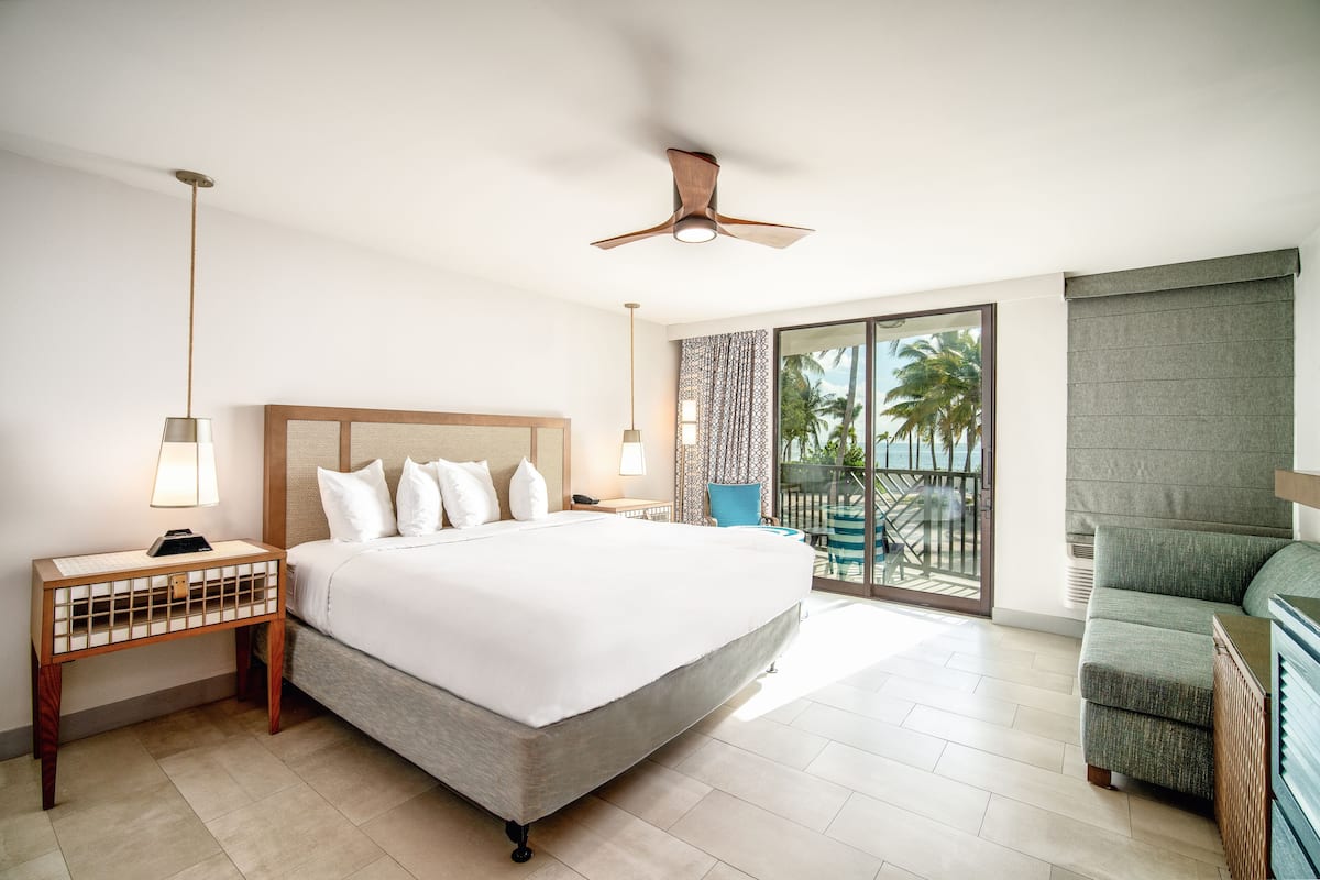 A hotel room with a large bed, a ceiling fan, a nightstand with a lamp, and a view of the outdoors through a sliding glass door.