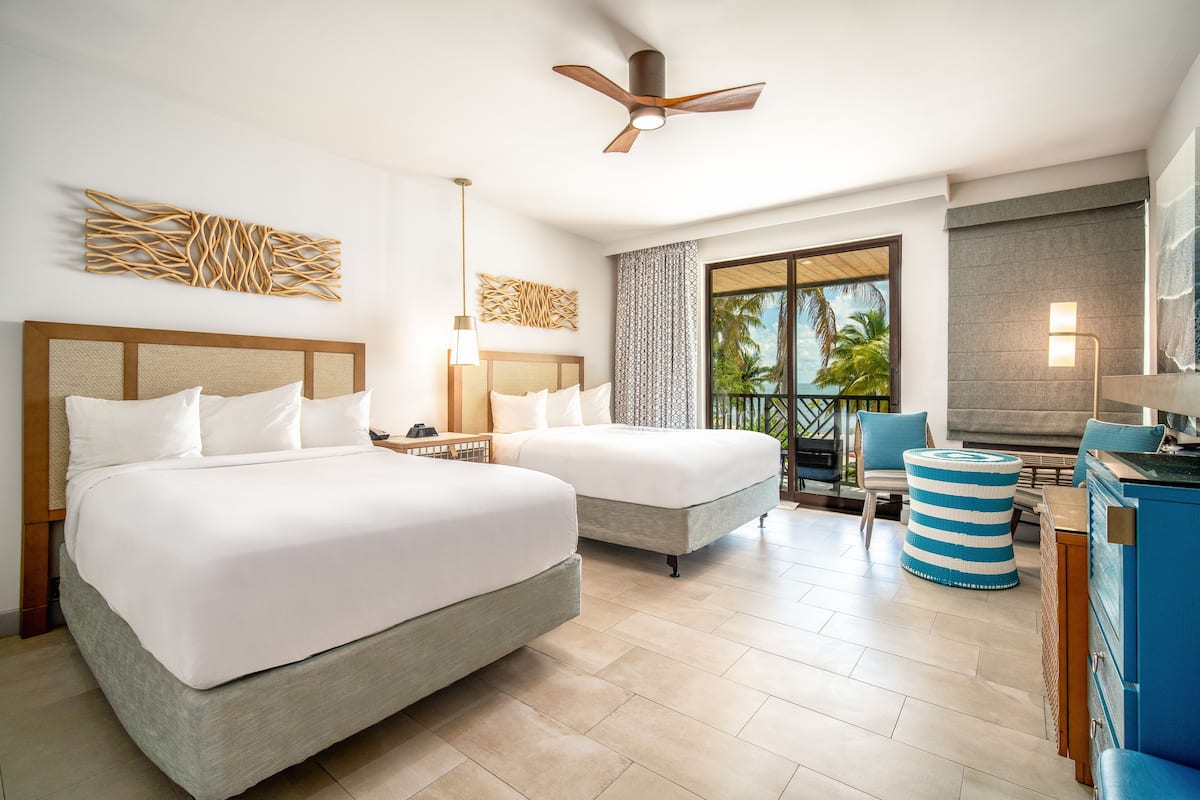A hotel room with two beds, a ceiling fan, a large window with a view of palm trees, and a balcony with a chair.