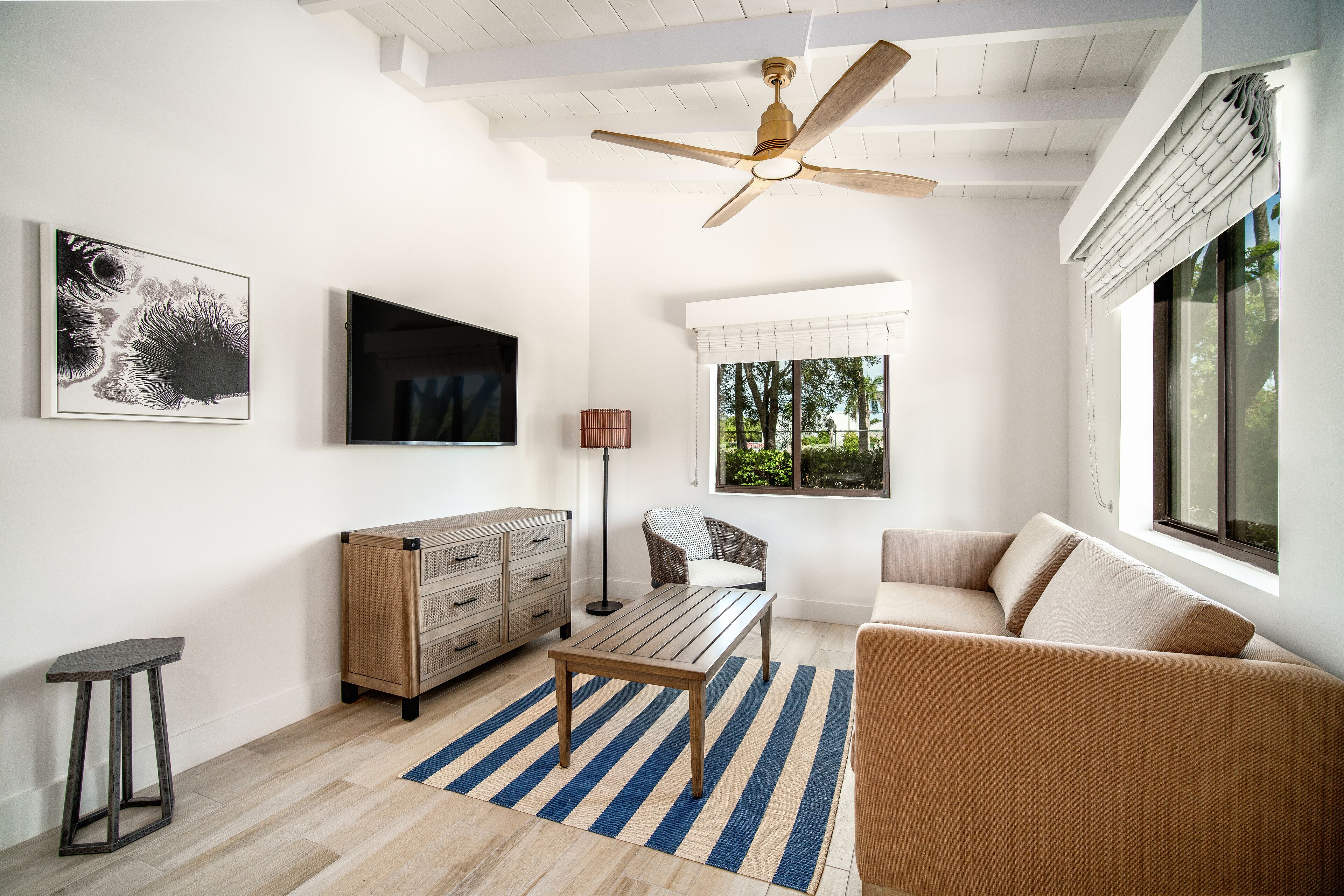 A modern living room with a ceiling fan, a flat-screen TV, a sofa, a coffee table, and a window with a view of trees.