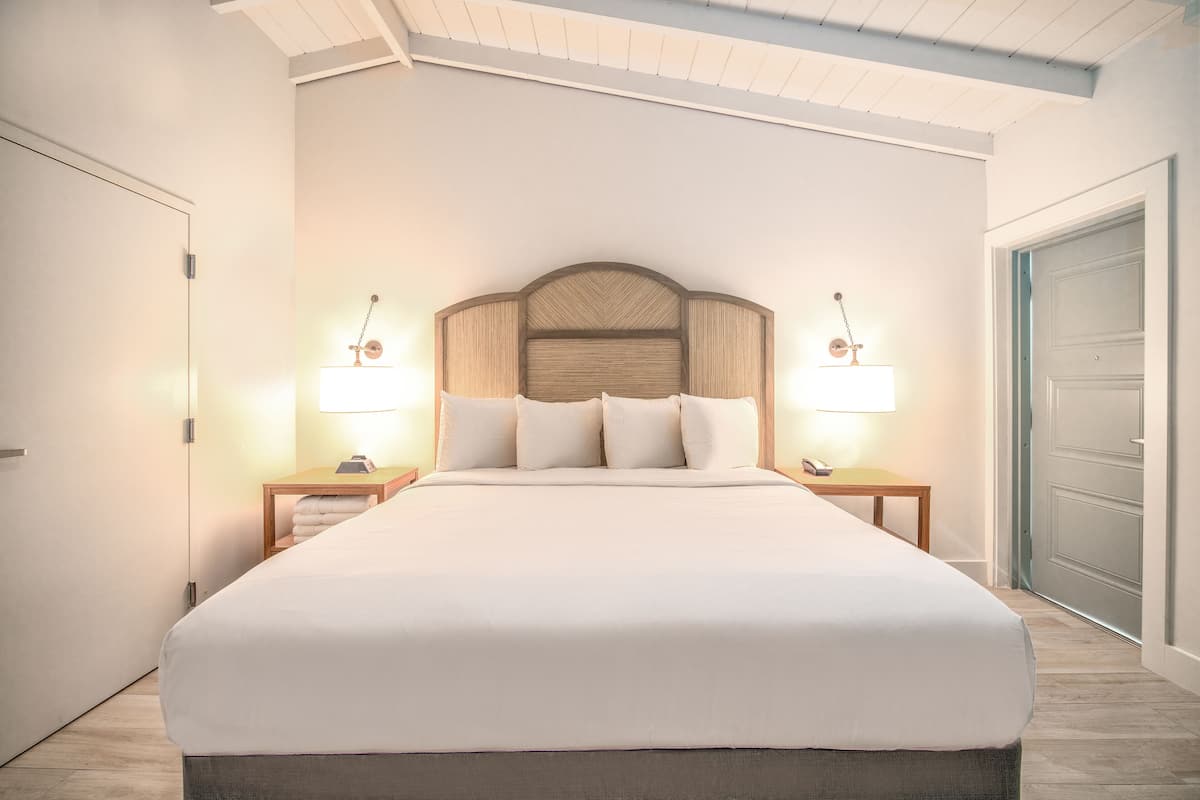 A neatly made bed in a hotel room with a wooden headboard, two bedside tables with lamps, and a door to the right.