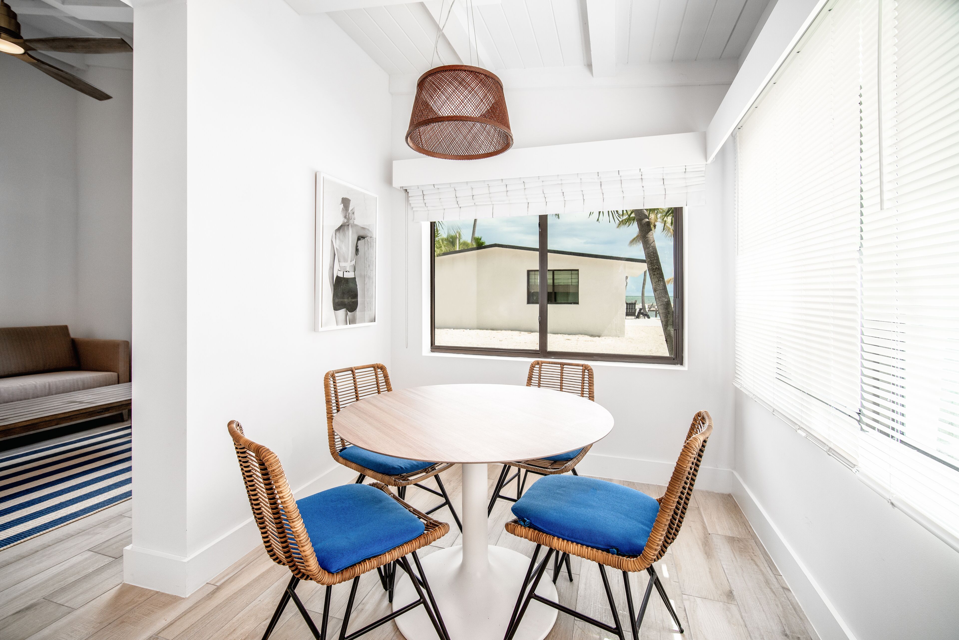 Un comedor con una mesa redonda y cuatro sillas de mimbre, un ventilador de techo y una ventana con persianas.