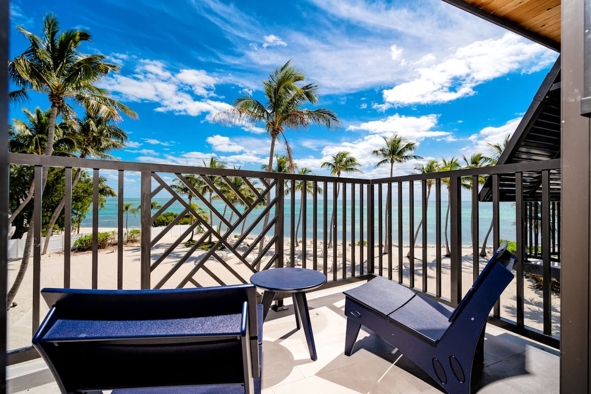 A balcony with two blue chairs and a small table, overlooking a beach with palm trees and the ocean.