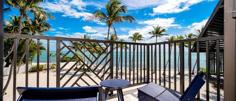 A balcony with two blue chairs and a small table, overlooking a beach with palm trees and the ocean.