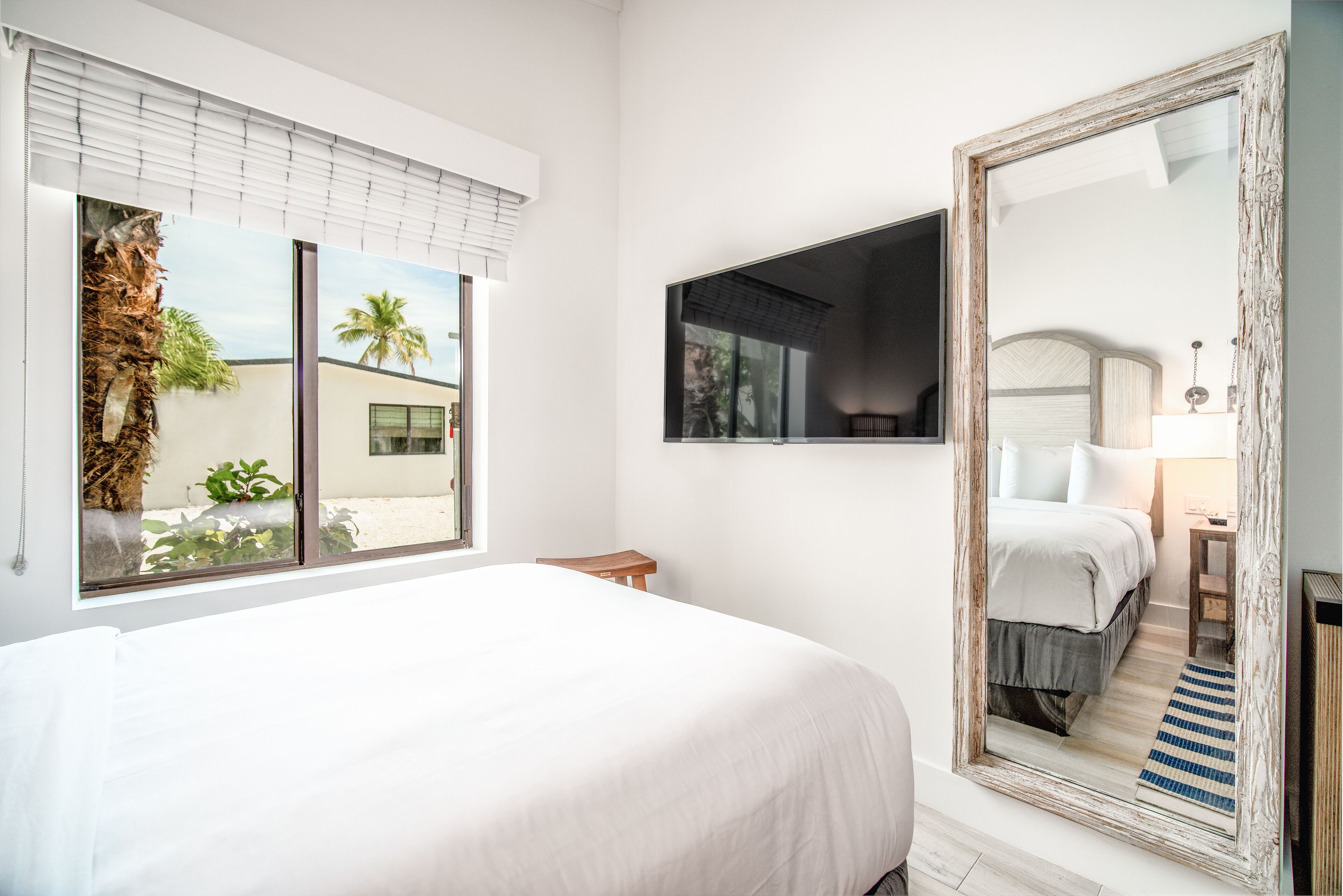 A bedroom with a large bed, a window with blinds, a mirror, and a view of palm trees and a building outside.