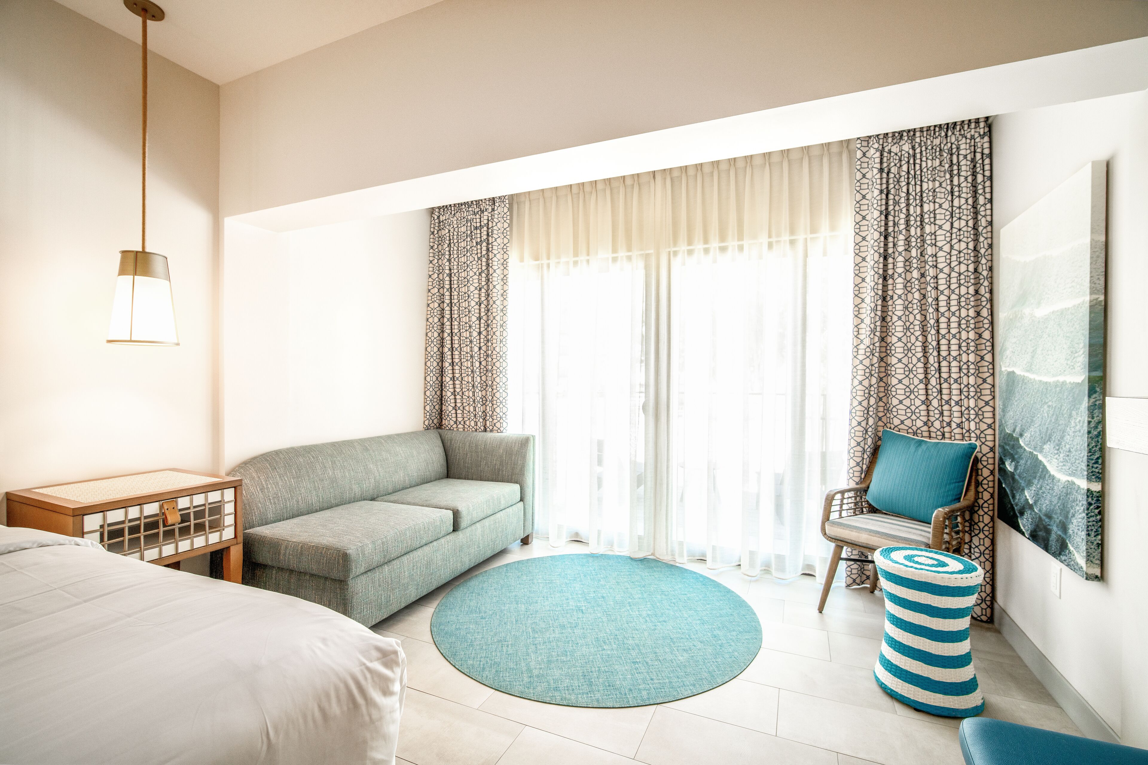 A hotel room with a bed, a sofa, a round rug, a chair with a cushion, a small table, and a hanging light fixture.