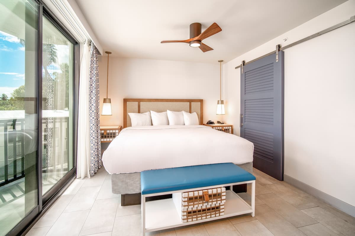 A modern bedroom with a large bed, a ceiling fan, bedside lamps, a bench, and a view through a sliding door.