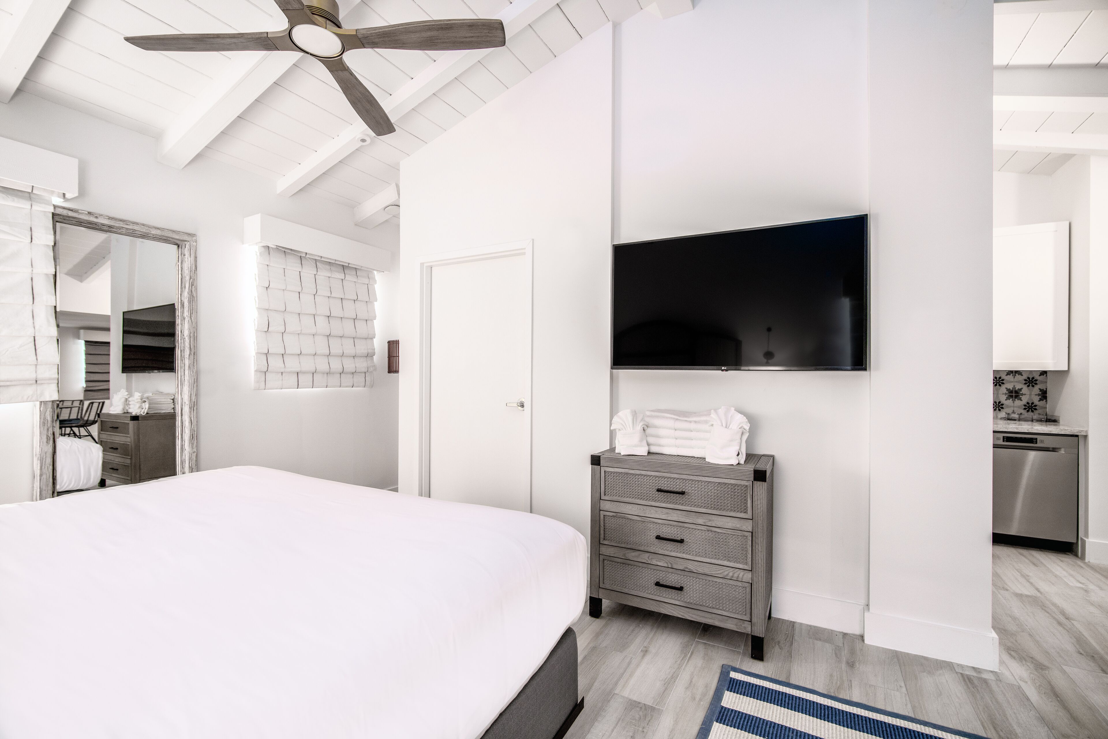 Ein modernes Schlafzimmer mit einem großen Bett, einem flachbildfernseher an der Wand, einem hölzernen Deckenventilator und einem durch eine offene Tür einsehbarer Küchenbereich.