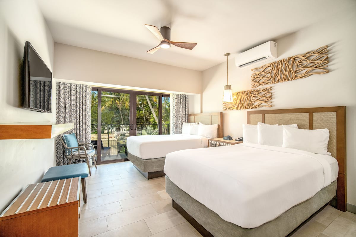 A hotel room with two beds, a ceiling fan, an air conditioning unit, a TV, a desk with a chair, and a view of greenery through the windows.