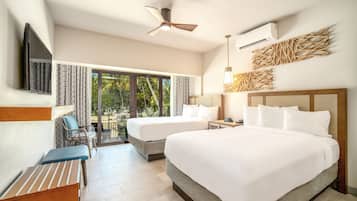 A hotel room with two beds, a ceiling fan, an air conditioning unit, a TV, a desk with a chair, and a view of greenery through the windows.