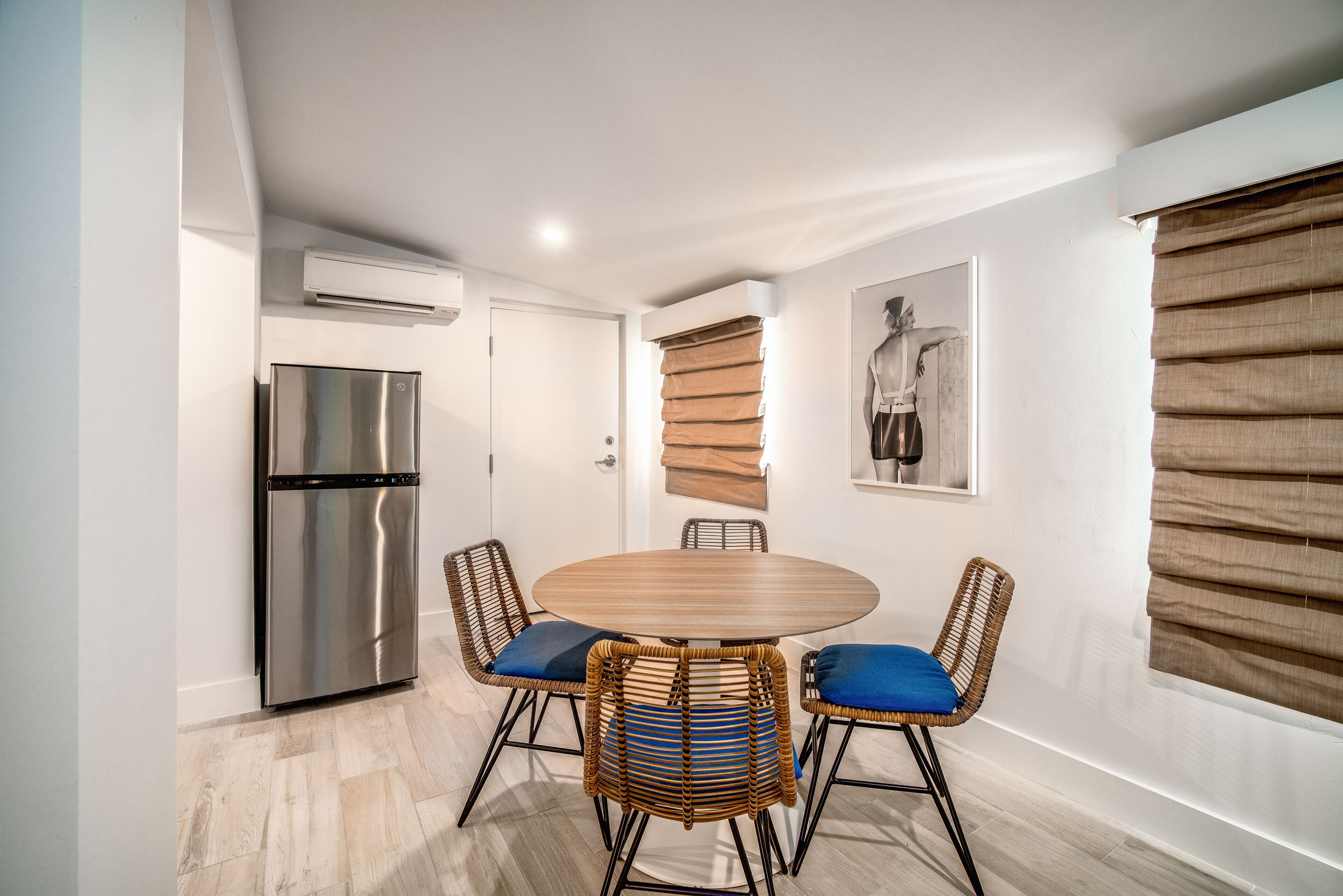 Una cocina moderna con un refrigerador de acero inoxidable, una mesa redonda de madera y sillas de mimbre, y un aire acondicionado instalado en la pared.