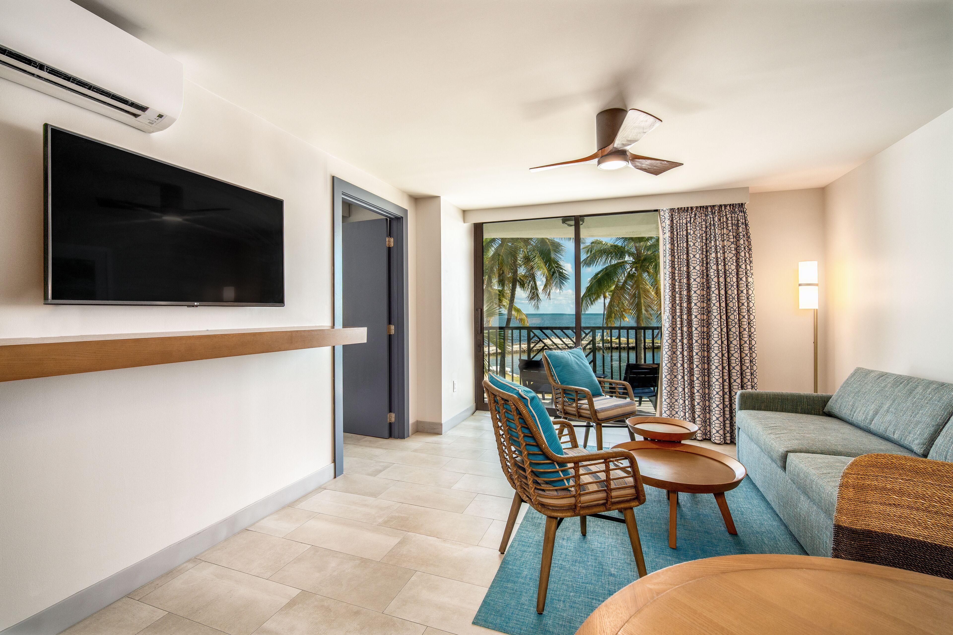 A modern living room with a flat-screen TV, a ceiling fan, a balcony with a view of the ocean, and a seating area with a sofa and chairs.