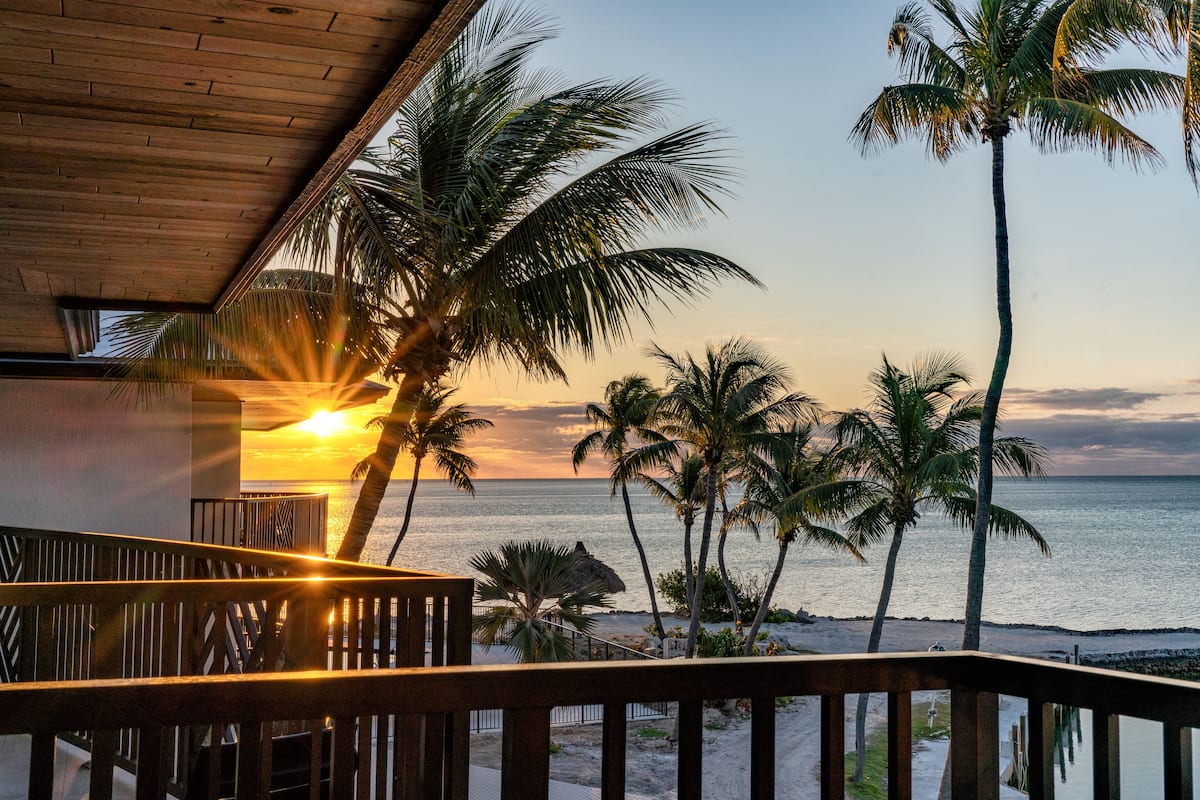 A wooden balcony overlooking a beach with palm trees and a sunset.