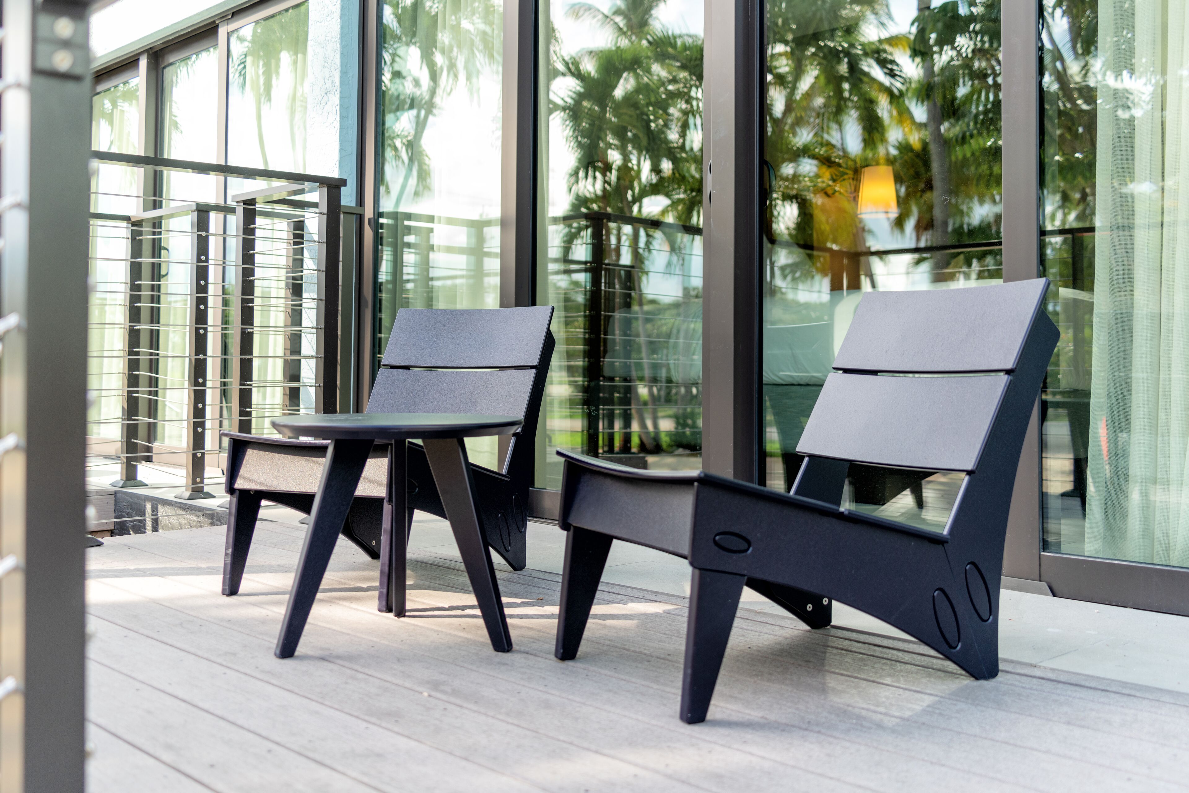 Two modern chairs and a small table on a balcony with large windows and a view of palm trees.