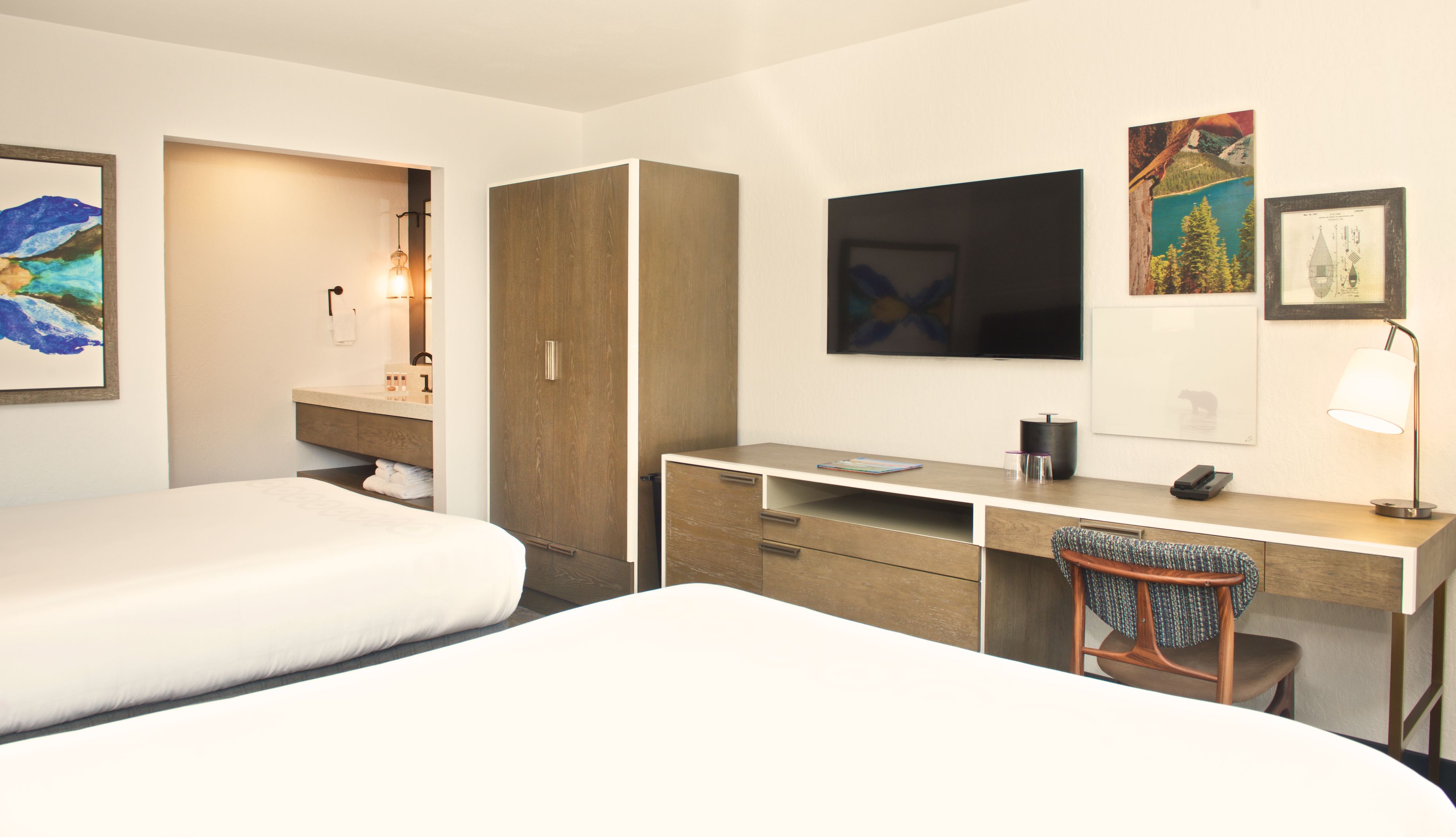 A hotel room with two beds, a desk, a chair, a TV, and artwork on the walls.