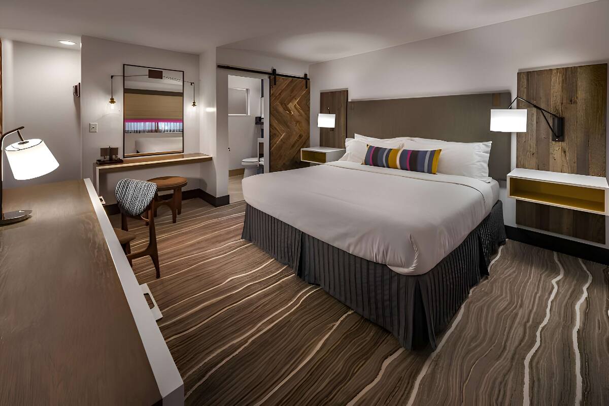 A hotel room with a large bed, a desk, a chair, a mirror, a wooden headboard, and a nightstand with a lamp.