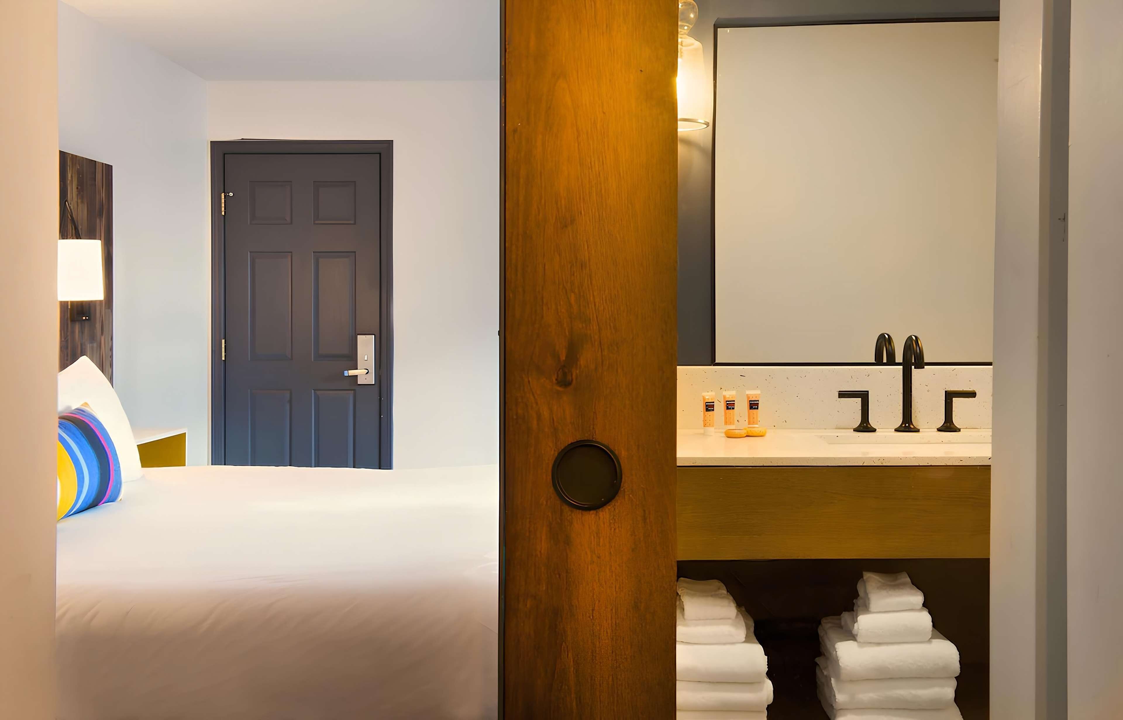 A hotel room with a bed, a wooden door, a bathroom with a sink and toiletries, and a mirror.
