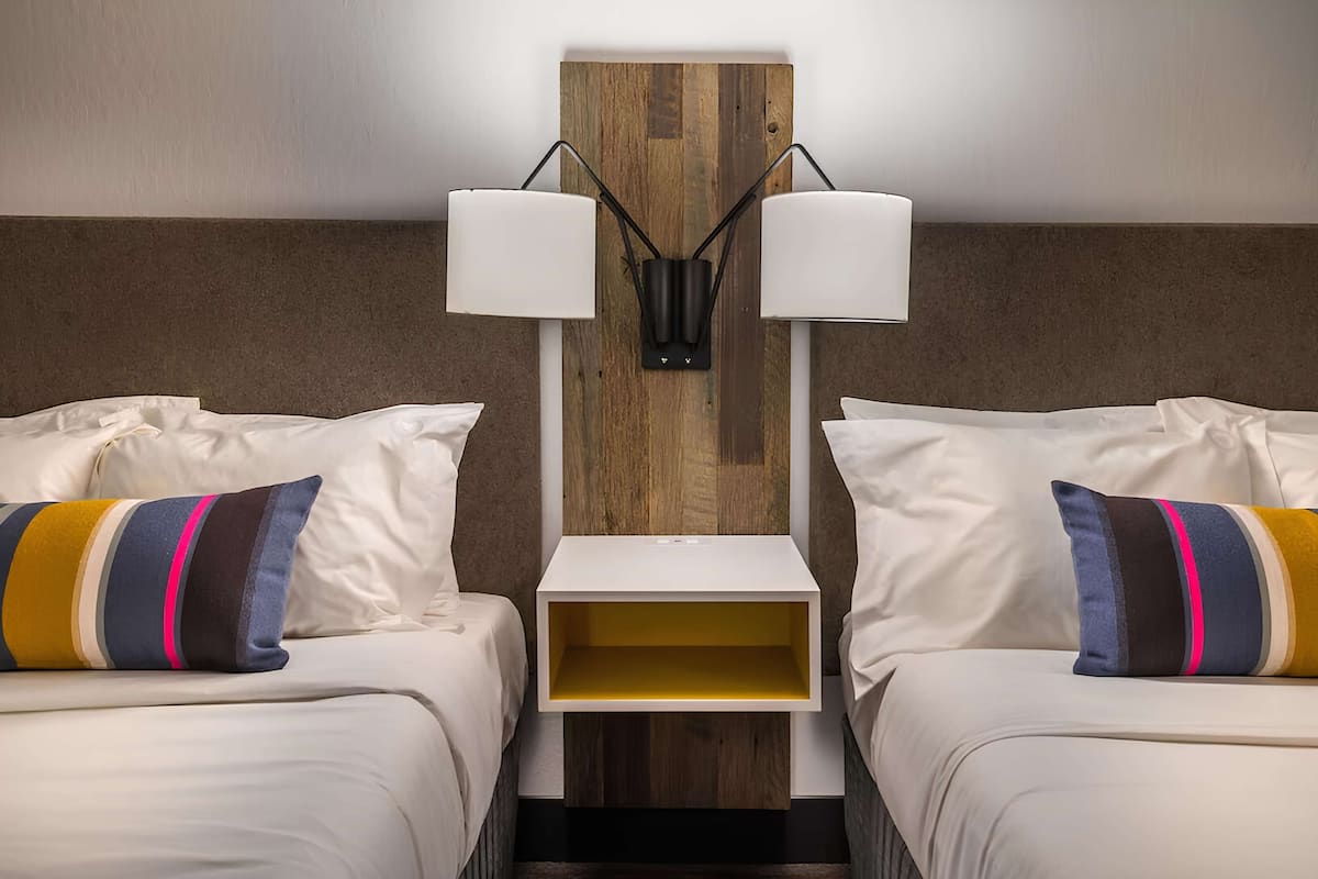 A hotel room with two beds, each with a pillow and a bedside table.