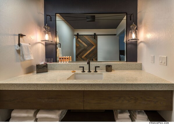 Un baño moderno con encimera de granito, un lavamanos y un espejo. Hay dos luces empotradas en la pared, un perchero y un mueble de tocador de madera.