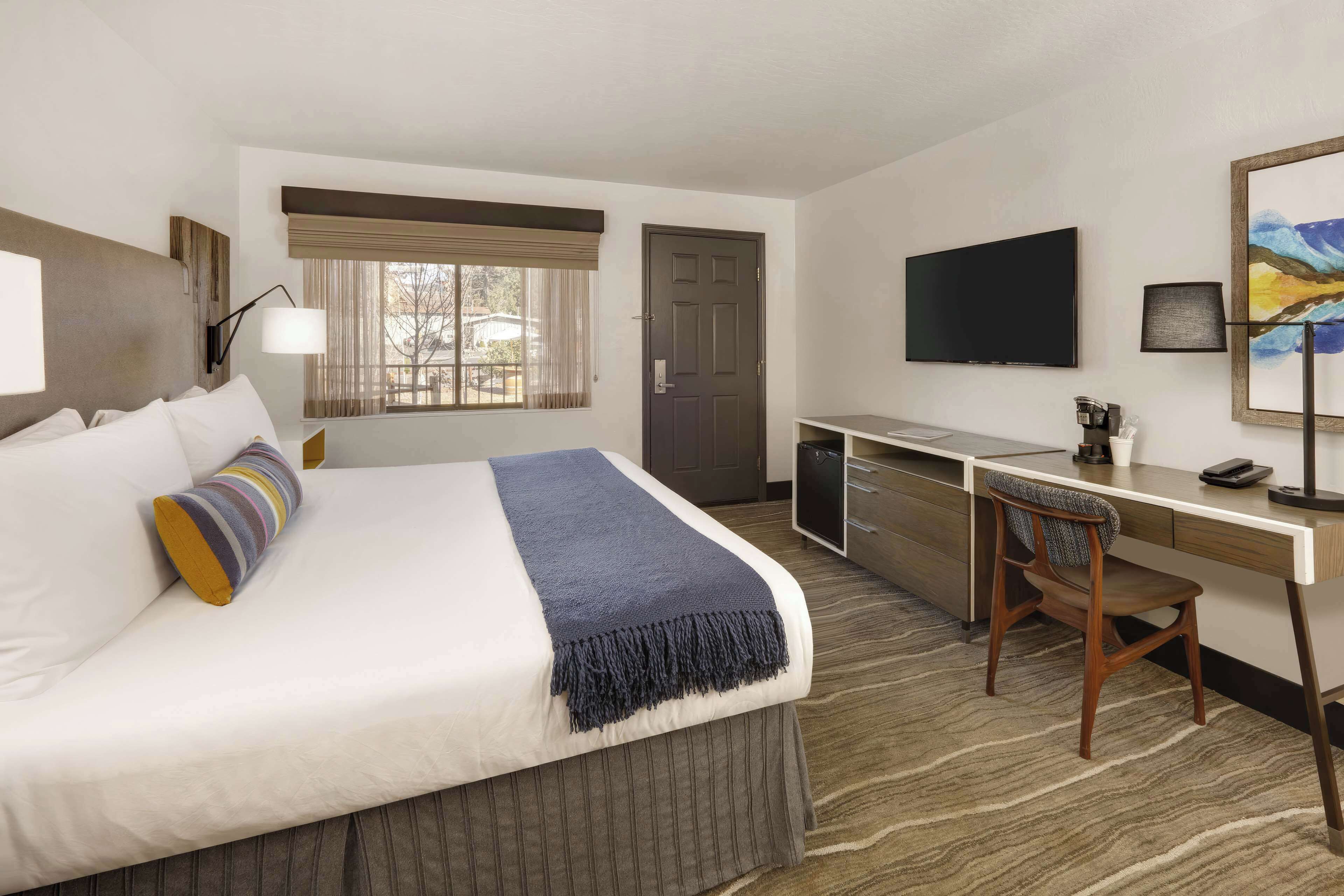 A hotel room with a bed, desk, chair, TV, and artwork.