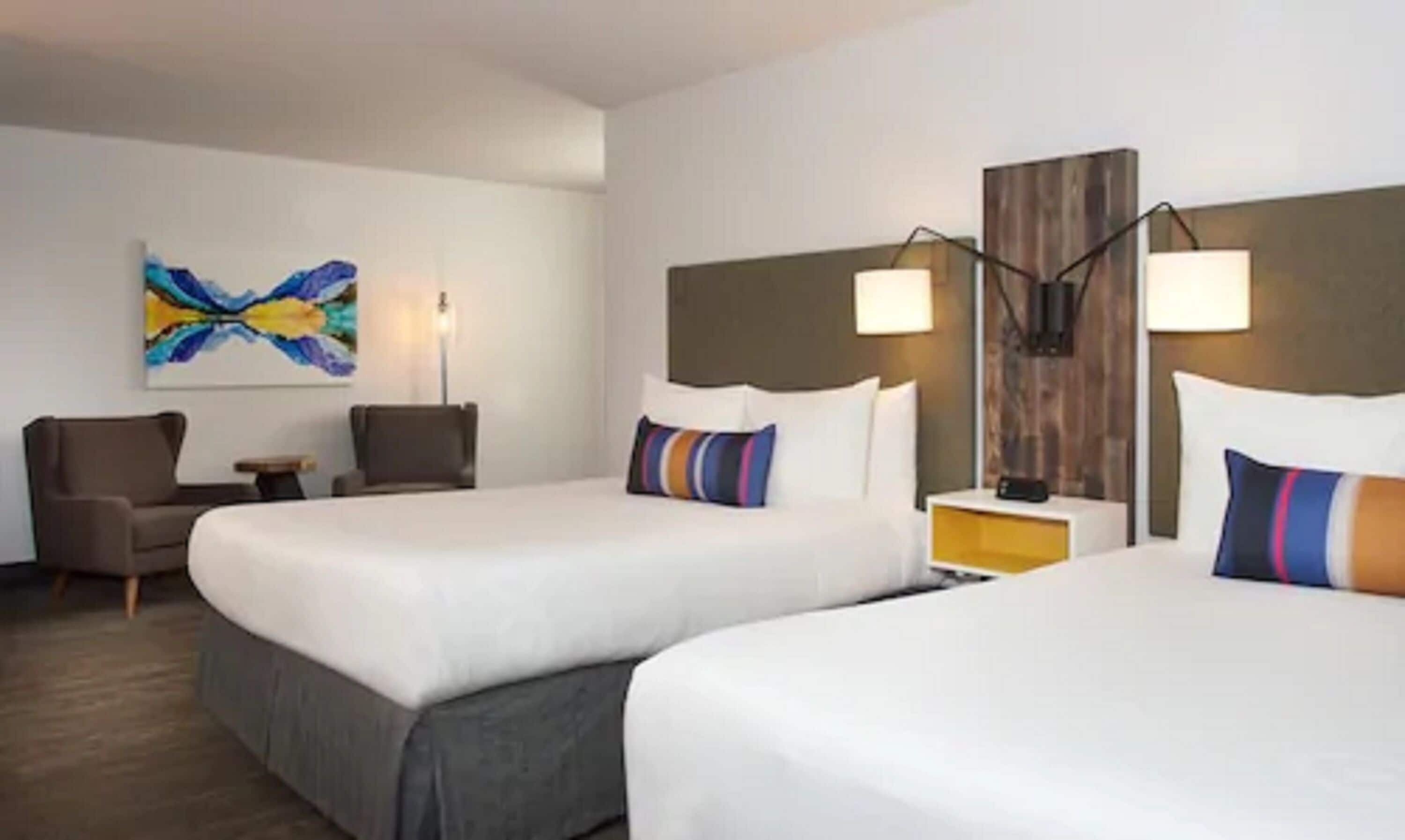 A hotel room with two beds, a desk, a chair, a lamp, and a painting on the wall.